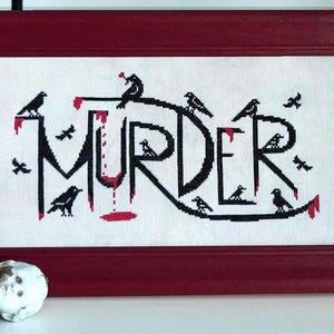 Op de afbeelding: Kruissteekkunstwerk in een bordeauxrode lijst. Het woord "MURDER" is in zwart gestikt met rode druppels en kraaien. Het kunstwerk staat op een witte achtergrond. Een vintage poppenhoofd en andere objecten staan op de voorgrond.