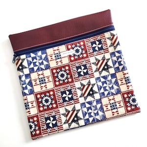 May include: A square zippered pouch featuring a patchwork quilt pattern in red, white, and blue with star and stripe motifs. The top section is made of textured dark maroon material, separated by a navy blue zipper. A small white label is visible.