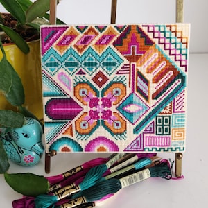 Może przedstawiać: Kolorowy wzór haftu krzyżykowego z geometrycznymi kształtami i kwiatowym wzorem w odcieniach różowego, turkusowego, pomarańczowego i białego. Haft krzyżykowy jest oprawiony i prezentowany na sztaludze.