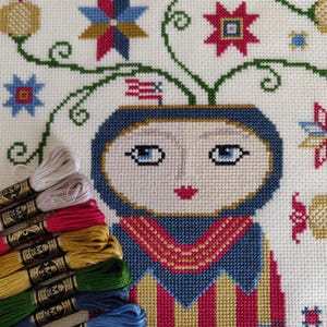 Puede incluir: Patrón de bordado de punto de cruz con un rostro estilizado con ojos azules, labios rojos y un vestido a rayas azules y amarillas. El rostro está rodeado de enredaderas verdes y flores de colores.