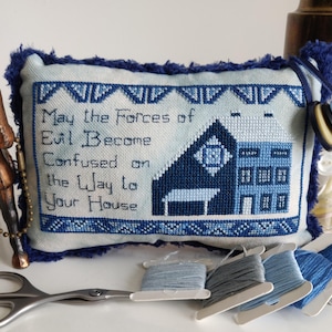 Może przedstawiać: Poduszka haftowana krzyżykiem w kolorze niebieskim i białym z tekstem "Niech siły zła zgubią się w drodze do twojego domu". Poduszka jest ozdobiona niebieskim domem i niebiesko-białą ramką.