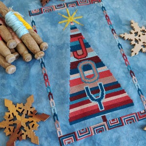 Może przedstawiać: Wzór haftu krzyżykowego przedstawiający choinkę z napisem "RADOŚĆ" napisanym kolorowymi paskami. Choinka jest otoczona niebiesko-białą ramką.