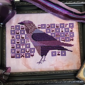 Puede incluir: Un bordado de punto de cruz morado y blanco de un cuervo con el alfabeto en cuadrados a su alrededor.