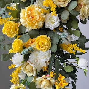May include: A cascading floral arrangement featuring yellow roses, peonies, and mimosa blossoms, accented with green eucalyptus leaves. The flowers are a mix of open and closed blooms, creating a textured and vibrant display.