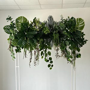 Puede incluir: Un arco de metal blanco adornado con exuberante vegetación artificial verde. El arreglo incluye grandes hojas de monstera, helechos y enredaderas colgantes, creando una estética tropical. Ideal para bodas o eventos.