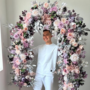 May include: A large floral archway with a variety of flowers in shades of pink, purple, and silver. The arch is decorated with roses, dahlias, and other flowers. The arch is a decorative element for weddings or events.