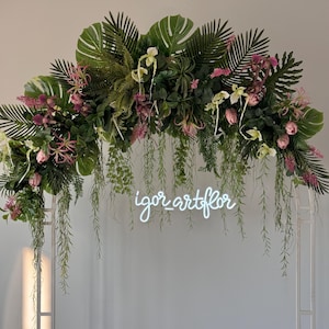 May include: A white metal arch decorated with lush greenery, including large monstera leaves, ferns, and pink and white flowers. The neon sign at the bottom reads "igor artflor". The arch is set against a plain white wall.