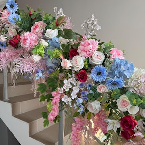 May include: A staircase railing adorned with a vibrant floral arrangement. The display features an array of flowers in shades of pink, white, blue, and red, including roses, peonies, and hydrangeas. Green foliage complements the colorful blooms, creating a lush, decorative display.