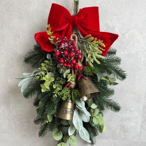 Könnte beinhalten: Ein festlicher Weihnachtsschmuck mit einer großen roten Samtschleife, künstlichem Grün, roten Beeren und zwei kleinen goldenen Glocken. Auf den Glocken stehen die Worte "SNOW" und "MONDAY". Ideal für festliche Weihnachtsdekorationen.