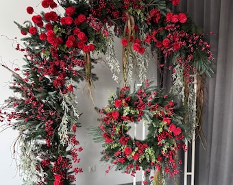 Puede incluir: Un arco de metal blanco decorado con una guirnalda roja y verde de ramas de pino, bayas rojas y pompones rojos. También se incluye una corona a juego.