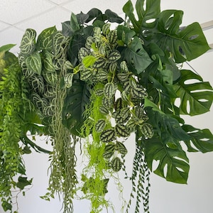 May include: An overhead display of various artificial plants in shades of green. Large, split-leaf Monstera leaves are interspersed with smaller, patterned leaves and trailing vines. The arrangement is lush and vibrant.