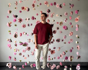 Puede incluir: Una persona está de pie frente a una pared decorada con numerosas flores rosas y blancas. La persona lleva una camiseta granate, pantalones blancos y zapatillas blancas. Las flores son de varios tamaños y tonos de rosa.