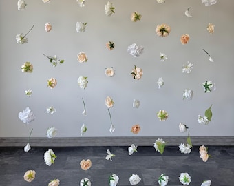 May include: An array of artificial flowers, including roses and other blossoms, suspended against a light gray wall. The flowers are in shades of white, cream, and peach, with green accents. The arrangement creates a floating floral display.