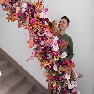 May include: A large, colourful floral arrangement with burgundy, pink, and orange flowers and foliage. The display includes roses, dahlias, and other blooms, creating a rich, vibrant cascade. Ideal for weddings or special occasions.