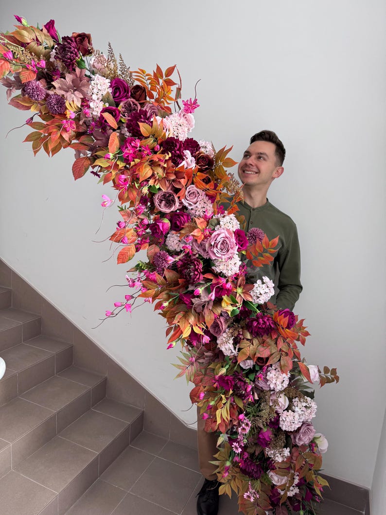 May include: A large floral arrangement with a cascade of burgundy, pink, and orange flowers and leaves. The display includes roses, dahlias, and other blooms, creating a rich and colourful arrangement. The flowers are arranged in a flowing, organic shape.