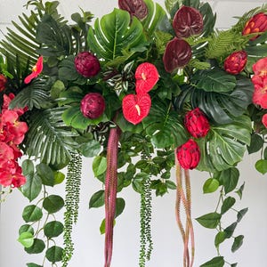 May include: An arrangement of artificial tropical flowers and foliage. The display features large green monstera leaves, red anthuriums, and cascading vines. Deep red, spherical flowers and long, trailing stems add to the vibrant composition.