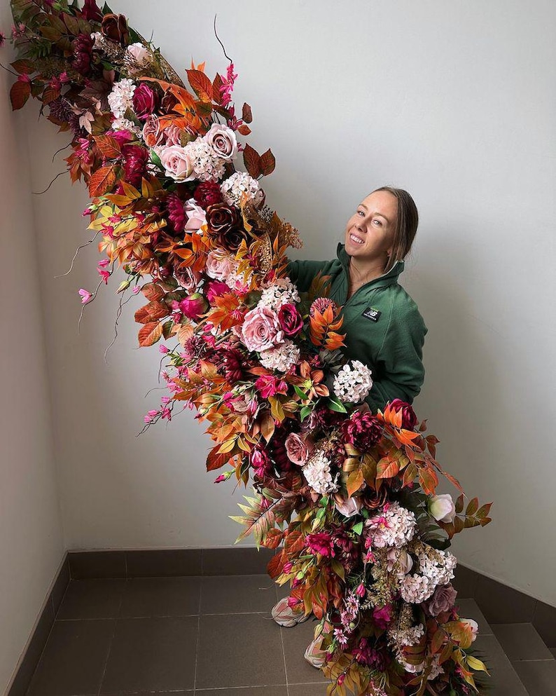 May include: A large floral arrangement with a cascade of colourful flowers and foliage. The arrangement includes roses, hydrangeas, and various leaves in shades of red, orange, pink, and white. The flowers are arranged in a cascading design.
