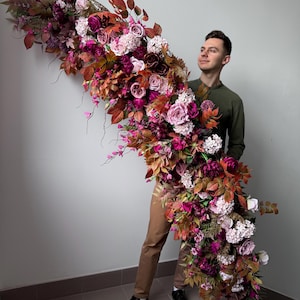 May include: A large, cascading floral arrangement with burgundy, pink, and orange flowers. The display includes roses, hydrangeas, and foliage, creating a full, textured look. The arrangement is held by a person.