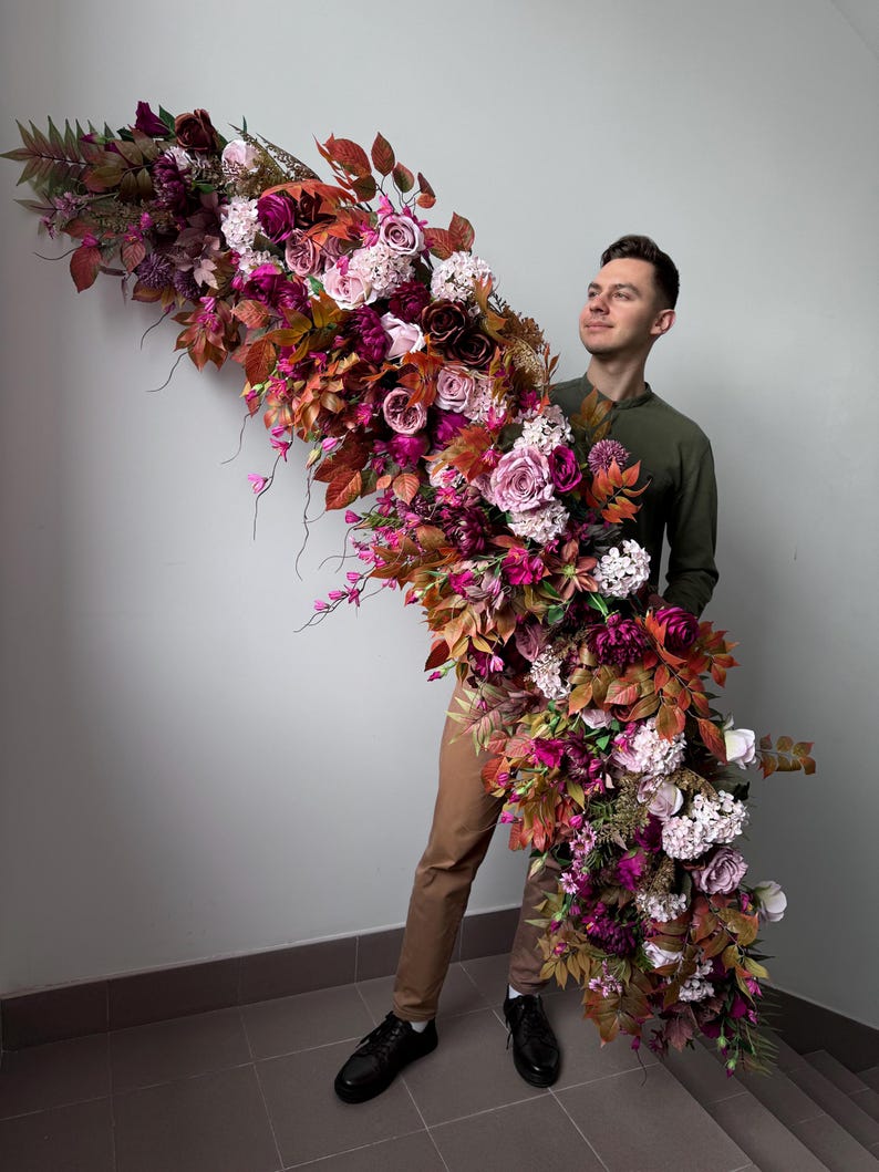 May include: A large, cascading floral arrangement with burgundy, pink, and orange flowers. The display includes roses, hydrangeas, and foliage, creating a full, textured look. The arrangement is held by a person.
