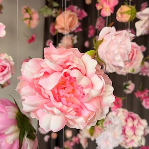 May include: A display of hanging artificial flowers in various shades of pink and white. The focal point is a large, fully bloomed peony with layered petals. Other flowers, including roses and smaller blooms, are suspended from above, creating a floral canopy.
