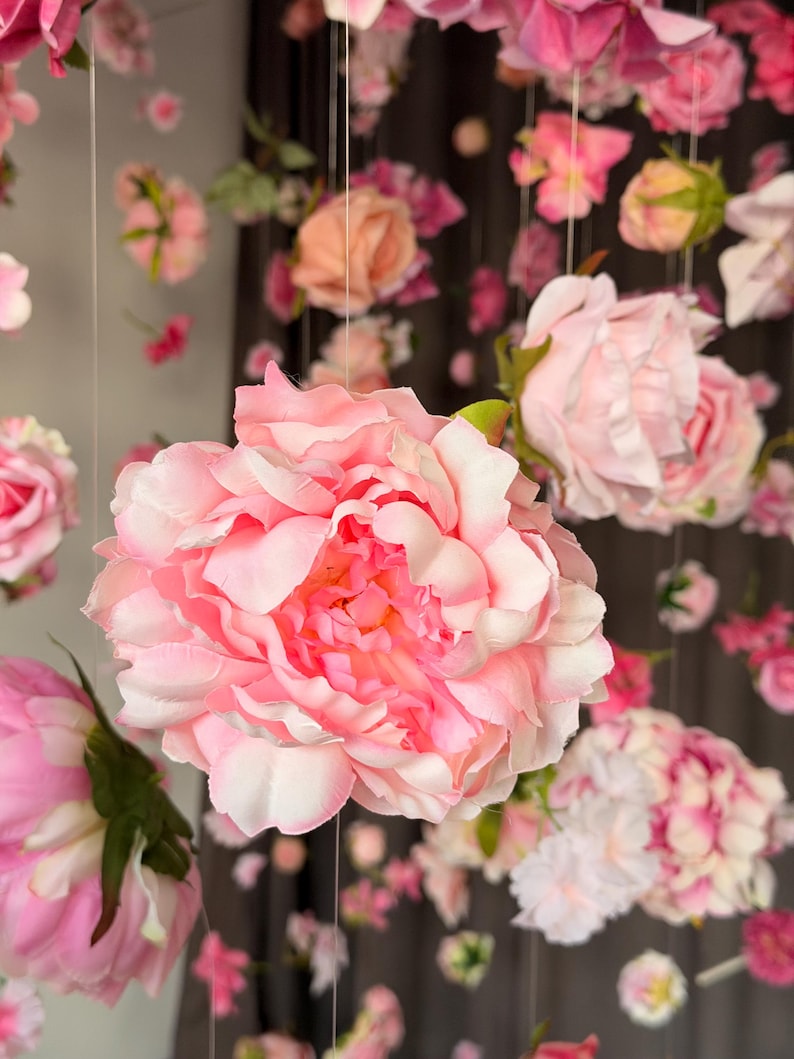 May include: A display of hanging artificial flowers in various shades of pink and white. The focal point is a large, fully bloomed peony with layered petals. Other flowers, including roses and smaller blooms, are suspended from above, creating a floral canopy.