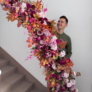 May include: A large, colourful floral arrangement with burgundy, pink, and orange flowers and foliage. The display includes roses, dahlias, and other blooms, creating a rich, vibrant cascade. Ideal for weddings or special occasions.