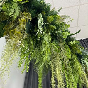 May include: An arrangement of various green artificial plants, including ferns, eucalyptus, and hanging vines. The foliage is a mix of light and dark greens, creating a layered and textured look. The arrangement is likely for decoration.
