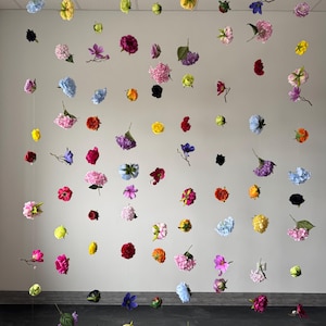 May include: A display of various colorful flowers suspended in the air against a neutral wall. The flowers, in shades of pink, yellow, blue, red, and purple, appear to be floating, creating a whimsical and artistic arrangement.