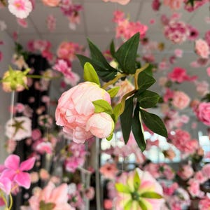 May include: A display of artificial flowers in various shades of pink and white, suspended from above. The arrangement includes peonies, roses, and other floral varieties, creating a soft, romantic aesthetic. Green leaves add contrast to the pastel colors.