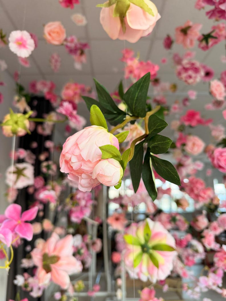 May include: A display of artificial flowers in various shades of pink and white, suspended from above. The arrangement includes peonies, roses, and other floral varieties, creating a soft, romantic aesthetic. Green leaves add contrast to the pastel colors.