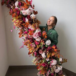 May include: A large floral arrangement with a cascade of colourful flowers and foliage. The arrangement includes roses, hydrangeas, and various leaves in shades of red, orange, pink, and white. The flowers are arranged in a cascading design.