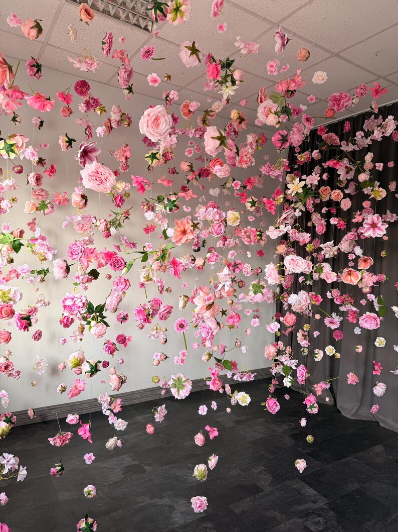 May include: A display of hanging pink and white flowers, including roses and other varieties, suspended from the ceiling. The flowers are arranged to create a visually appealing, immersive floral installation. The background is a neutral color.