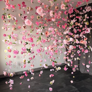 May include: A display of hanging pink and white flowers, including roses and other blossoms, suspended from the ceiling. The flowers are arranged to create a visually appealing, floral installation. The background is a neutral wall and dark floor.