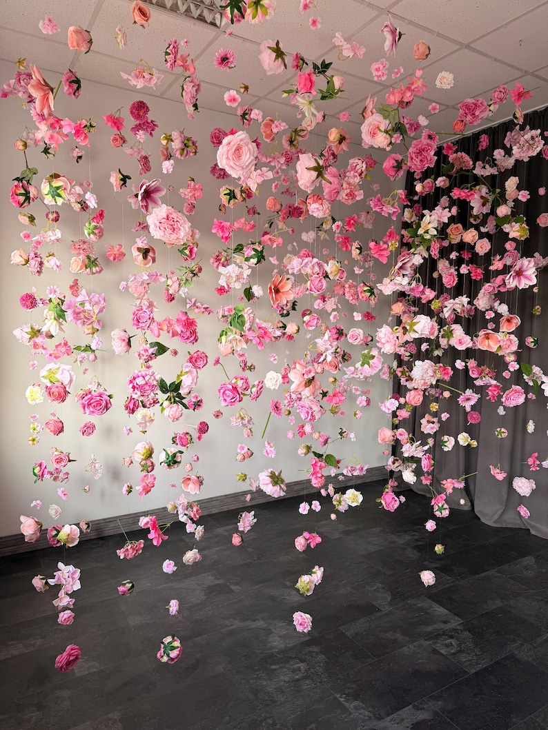 May include: A display of hanging pink and white flowers, including roses and other blossoms, suspended from the ceiling. The flowers are arranged to create a visually appealing, floral installation. The background is a neutral wall and dark floor.