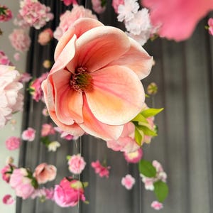 May include: A close-up of a large, peach-colored flower with multiple petals, suspended from a string. The flower is surrounded by other smaller pink and white flowers, creating a floral display. The background is a dark gray curtain.