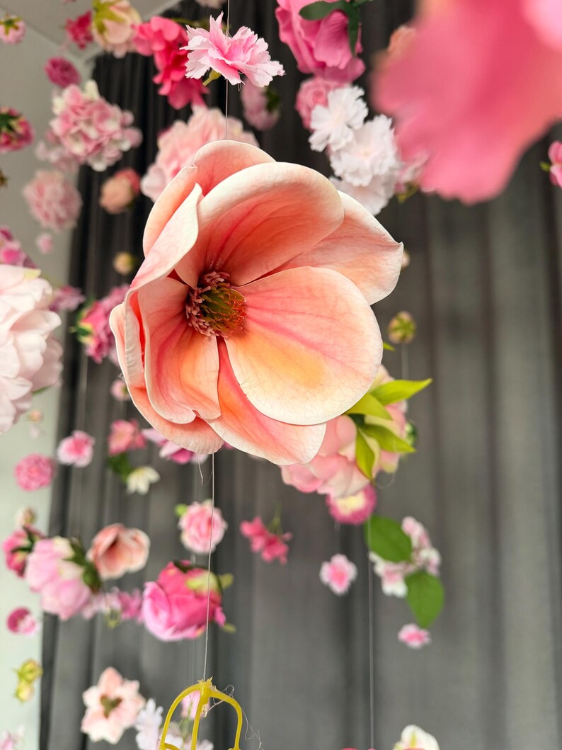 May include: A close-up of a large, peach-colored flower with multiple petals, suspended from a string. The flower is surrounded by other smaller pink and white flowers, creating a floral display. The background is a dark gray curtain.