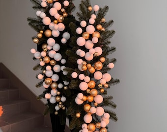 Op de afbeelding: Twee kunstmatige kerstbomen versierd met witte en gouden ballen. De bomen zijn verticaal gerangschikt, met de ornamenten geclusterd langs de takken. De bomen staan tegen een neutrale achtergrond, met een trap zichtbaar in het onderste deel van de afbeelding.