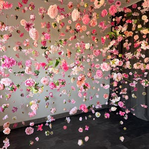 May include: A display of hanging pink and white flowers, creating a floral curtain effect. Various flower types and shades of pink are suspended from the ceiling, with some flowers scattered on the dark floor.