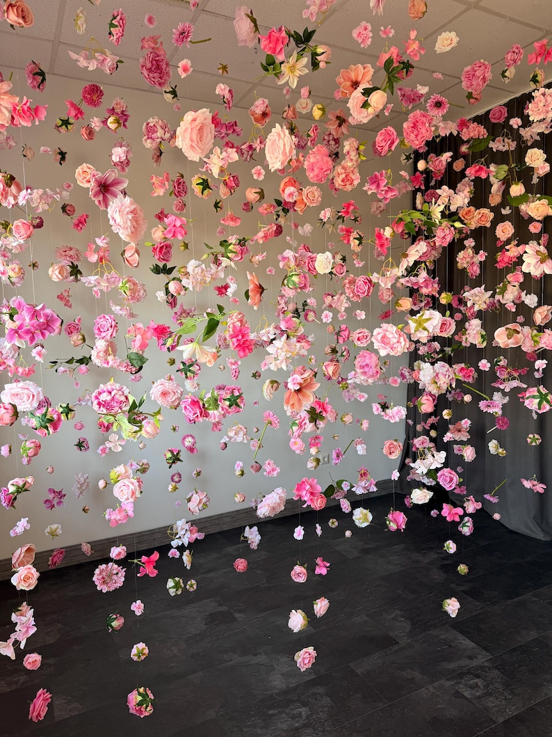 May include: A display of hanging pink and white flowers, creating a floral curtain effect. Various flower types and shades of pink are suspended from the ceiling, with some flowers scattered on the dark floor.
