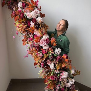 Op de afbeelding: Een groot bloemstuk met een waterval van kleurrijke bloemen en bladeren. Het arrangement bevat rozen, hortensia's en diverse bladeren in tinten roze, rood, oranje en bruin. De bloemen zijn in een watervalstijl gerangschikt.