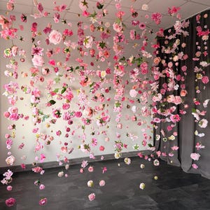 May include: A room decorated with hanging pink and white flowers. The flowers are suspended from the ceiling, creating a floral curtain effect. The flowers are of various sizes and types, adding depth and texture to the display. The floor is dark, contrasting with the light colors of the flowers.
