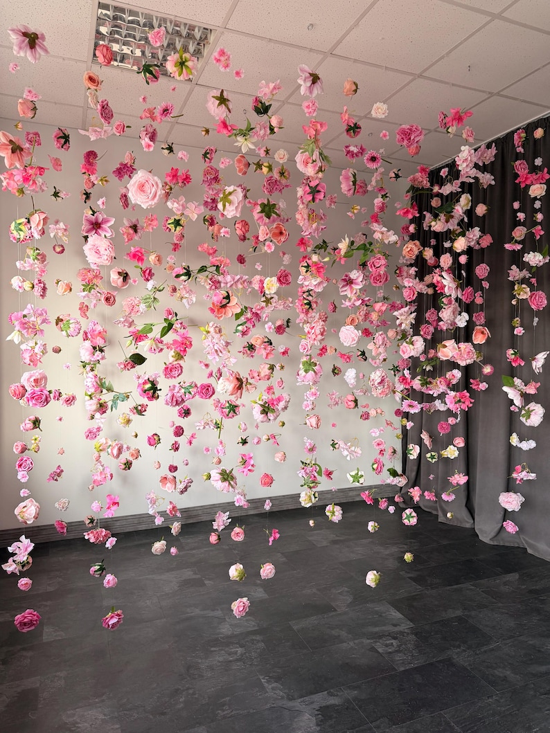 May include: A room decorated with hanging pink and white flowers. The flowers are suspended from the ceiling, creating a floral curtain effect. The flowers are of various sizes and types, adding depth and texture to the display. The floor is dark, contrasting with the light colors of the flowers.