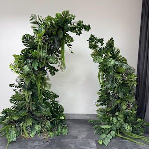 May include: A vibrant green archway constructed from artificial tropical foliage. The arch features a variety of leaf types, including large monstera leaves and smaller ferns. The arch is a vivid green and is positioned against a white wall.