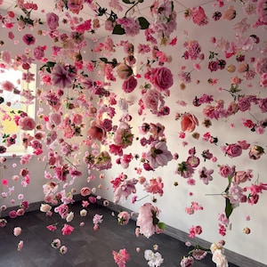 May include: A room filled with hanging pink and white flowers, creating a floral canopy. Various types of blooms, including roses and peonies, are suspended from the ceiling, with some scattered on the floor. The walls are white, and the floor is dark wood.