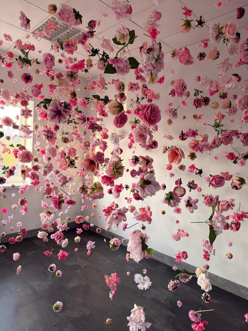 May include: A room filled with hanging pink and white flowers, creating a floral canopy. Various types of blooms, including roses and peonies, are suspended from the ceiling, with some scattered on the floor. The walls are white, and the floor is dark wood.