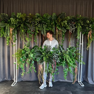 以下が含まれることがあります： 緑豊かな熱帯植物と緑で飾られた白い金属製のアーチ。アーチは灰色のカーテンの前に立っています。