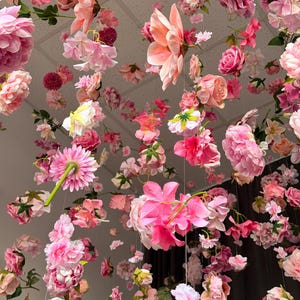 May include: An overhead view of a display of various pink and peach-colored flowers suspended from the ceiling. The arrangement includes roses, peonies, and other floral varieties, creating a soft and romantic aesthetic. The flowers are of different sizes and shapes.