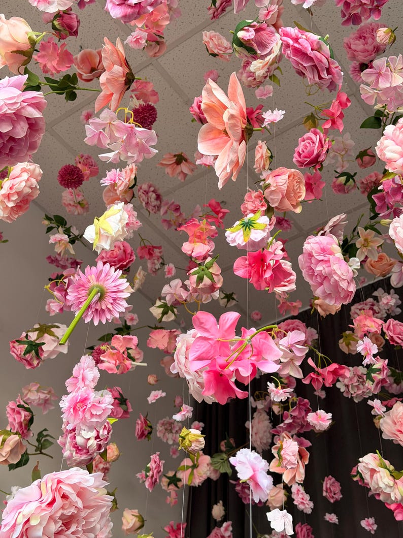 May include: An overhead view of a display of various pink and peach-colored flowers suspended from the ceiling. The arrangement includes roses, peonies, and other floral varieties, creating a soft and romantic aesthetic. The flowers are of different sizes and shapes.