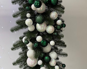 Op de afbeelding: Een kerstslinger versierd met groene en witte ballen. De slinger heeft een mix van matte en iriserende witte bollen, afgewisseld met glanzende en getextureerde groene ballen. De groenblijvende takken vormen een feestelijke achtergrond.