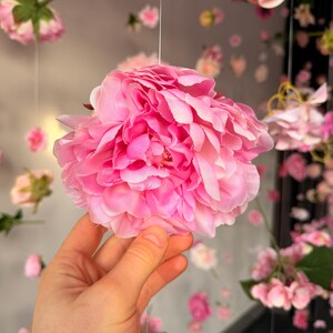 May include: A large, vibrant pink peony flower with layered petals is held in a hand. The background features a soft focus of other pink flowers, creating a floral theme.